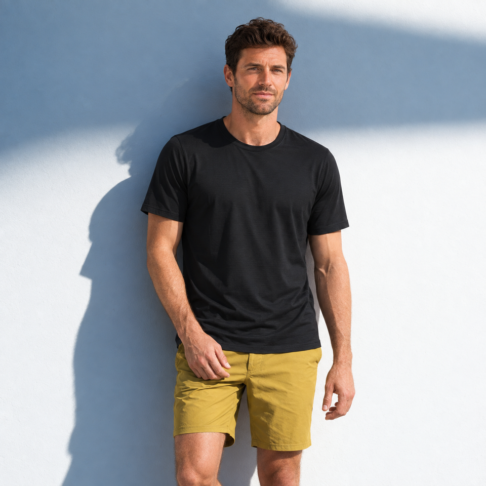 Mann in schwarzem Montevaro-T-Shirt und senffarbenen Shorts vor einer hellen Wand